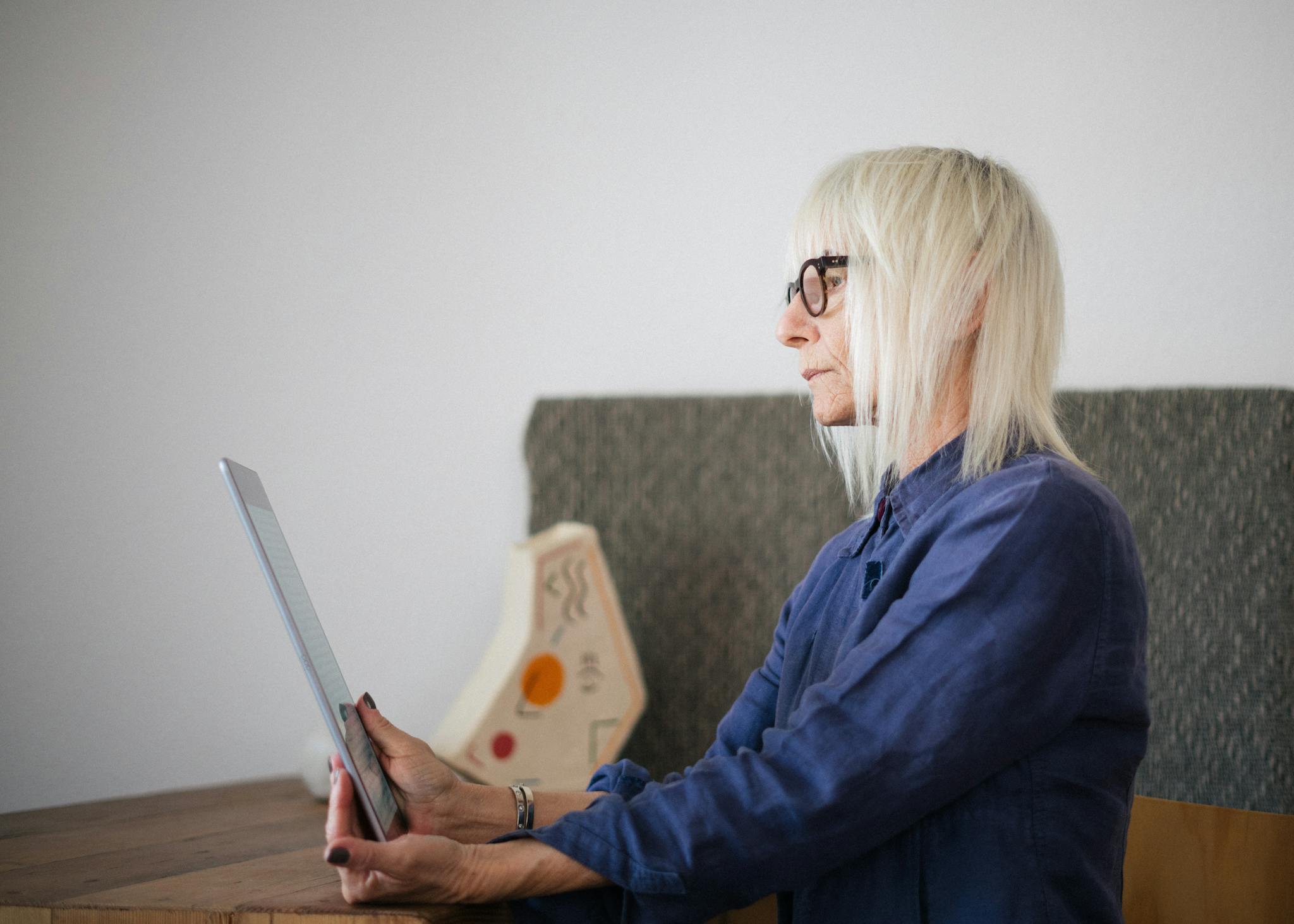 Side view of stylish concentrated aged woman in casual outfit sitting with tablet at table at home